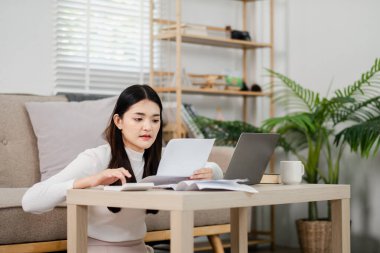 Bir kadın elinde dizüstü bilgisayar ve bir yığın kağıtla masada oturuyor. Kağıtlara bakıyor ve işine odaklanmış gibi görünüyor. Odanın rahat ve rahat bir atmosferi var.