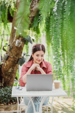 Önünde dizüstü bilgisayarı olan bir kadın masada oturuyor. O kırmızı bir gömlek giyiyor ve ellerini çapraz yaptırıyor. Sahne rahatlamış ve rahat bir atmosferi işaret ediyor, muhtemelen bir parkta ya da kafede.