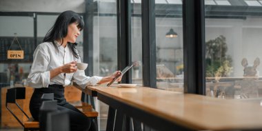 Bir kadın bir kafede bir masada oturuyor, tabletine bakıyor ve kahve içiyor. Dinlenme ve rahatlama kavramı, kadın dinlenirken kahve ve tabletinin tadını çıkarıyor.