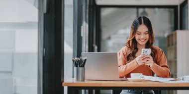 Bir kadın dizüstü bilgisayarı ve cep telefonu olan bir masada oturuyor. Gülümsüyor ve eğleniyor.