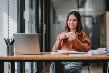 Bir kadın elinde dizüstü bilgisayar ve bir fincan kahveyle masada oturuyor. Gülümsüyor ve kahvesinin tadını çıkarıyor.
