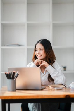 Bir kadın dizüstü bilgisayarı ve kalemi olan bir masada oturuyor. Gülümsüyor ve mutlu.
