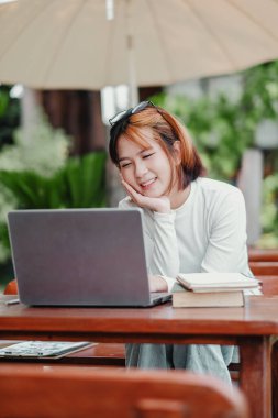 Bir kadın elinde dizüstü bilgisayar ve kitapla masada oturuyor. Gülümsüyor ve eğleniyor.