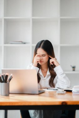 Masasında dizüstü bilgisayarı ve not defteri olan bir kadın oturuyor. Laptop ekranına bakıyor ve gözlerini ovalıyor. Sahne onun çalıştığını ya da çalıştığını ve yorgun ya da stresli olduğunu gösteriyor.