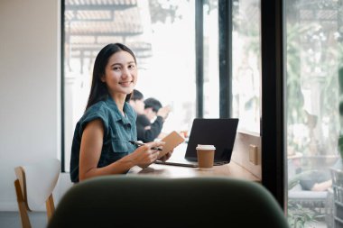Bir kadın dizüstü bilgisayarı ve not defteriyle masada oturuyor. Gülümsüyor ve eğleniyor. Sahne rahat ve rahat bir atmosferi çağrıştırıyor. Kadın çalışıyor olabilir.