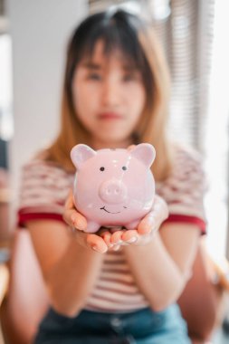 Bir kadının elinde pembe bir kumbara var. O, çizgili bir gömlek giyiyor. Domuz kumbarası pembe ve üzerinde gülen bir yüz var.