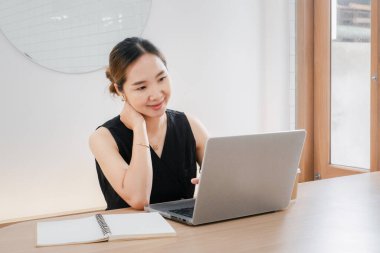 Masasında dizüstü bilgisayarı ve not defteri olan bir kadın oturuyor. Gülümsüyor ve işinden zevk alıyor.