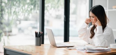 Bir kadın elinde dizüstü bilgisayar ve bir fincan kahveyle masada oturuyor. O, beyaz bir gömlek giyiyor ve hayal kırıklığı içinde.