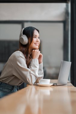 Kulaklık takan bir kadın elinde laptop ve bir fincan kahveyle bir masada oturuyor. Gülümsüyor ve eğleniyor.