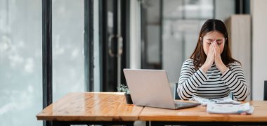 Bir kadın elinde dizüstü bilgisayar ve bir yığın kağıtla masada oturuyor. Dizüstü bilgisayara bakıyor ve zor bir durumda.