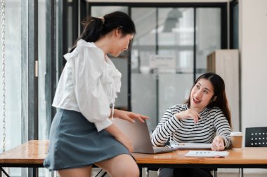 İki kadın bir masada oturuyor, biri dizüstü bilgisayarı işaret ediyor. İki kadın bir projede birlikte çalıştığı için sahne dostane ve işbirlikçi.