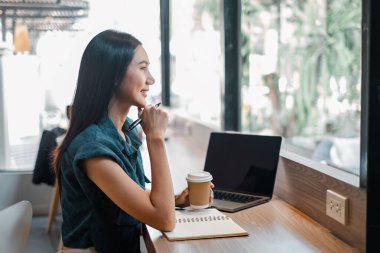 Bir kadın elinde dizüstü bilgisayar ve bir fincan kahveyle masada oturuyor. Gülümsüyor ve düşünüyor.