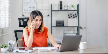 Turuncu gömlekli bir kadın dizüstü bilgisayarı ve kalemi olan bir masada oturuyor. Bir şey düşünüyor ya da düşünüyor.