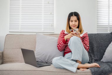 Bir kadın elinde cep telefonu ve dizüstü bilgisayarla kanepede oturuyor. Çoklu görev kavramı ve teknolojiye bağlı olmak