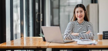 Bir kadın elinde dizüstü bilgisayar ve bir fincan kahveyle masada oturuyor. Gülümsüyor ve işinden zevk alıyor.