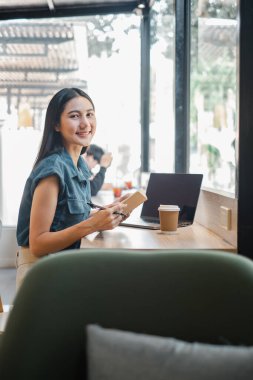 Bir kadın elinde dizüstü bilgisayar ve kitapla masada oturuyor. Gülümsüyor ve eğleniyor.