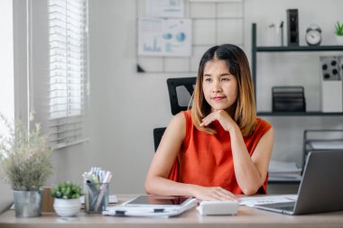 Modern bir ev ofisinde genç bir kadın. Bilgisayarı ve belgeleri olan bir masada düşünüyor ve çalışıyor..