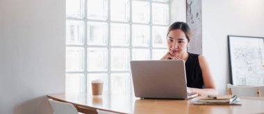 Modern bir ofis masasında dizüstü bilgisayarda çalışan genç bir kadın. Elinde kahve fincanı ve not defteri var. Cam bir pencereden ışıkla aydınlatılıyor..