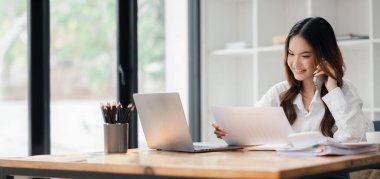 Ev ofisinden çalışan genç bir kadın, bilgisayarını ve belgelerini kullanıyor, telefonda gülümsüyor ve konuşuyor..