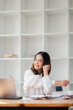 Modern bir ofiste masasında gülümseyen ve gerinen beyaz gömlekli genç bir kadın. Bir dizüstü bilgisayar ve kitaplar ahşap masanın üzerinde, arka planda beyaz raflar var..