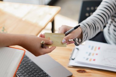 Arka planda dizüstü bilgisayar ve defter olan ahşap bir masanın üzerinde kredi kartı alışverişinde bulunan ellerin yakın çekimi..