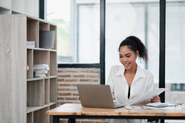 Beyaz gömlekli gülümseyen genç bir kadın, tuğla duvarlı ve geniş pencereli modern bir ofiste, ahşap bir masanın üstünde çalışıyor..
