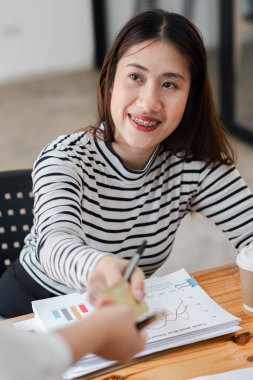 Diş telleriyle gülümseyen ve modern ofis ortamında kartvizitini teslim eden genç bir kadın. Evraklar ve kahve fincanı masanın üstünde..