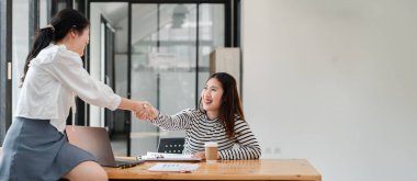 İş ortamında tokalaşan iki kadın. Bir kadın beyaz gömlek giyiyor ve diğeri siyah beyaz çizgili gömlek giyiyor.