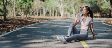 Spor kıyafetli bir kadın parkta otururken su içiyor, spor salonunu ve açık hava egzersizlerini renklendiriyor..