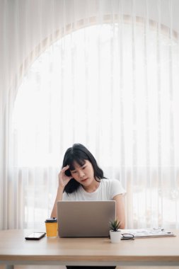 Ev ofisinin masasında dizüstü bilgisayarı üzerinde çalışan genç bir kadın. Elinde kahve ve belgelerle büyük bir penceresi ve perdesi olan parlak bir odada..