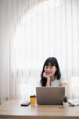 Ev ofisinin masasında dizüstü bilgisayarda çalışan genç bir kadın kahve ve akıllı telefonuyla perdelerin arasından geçen parlak doğal ışık..