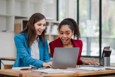 İki kadın önlerinde bir dizüstü bilgisayarla bir masada oturuyorlar. Gülümsüyorlar ve iyi vakit geçiriyor gibi görünüyorlar.