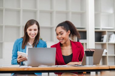 Modern bir ofiste birlikte çalışan iki profesyonel kadın, dizüstü bilgisayarlar ve dijital cihazlar kullanarak, gülümseyerek ve işbirliği yaparak.