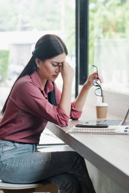 Genç bir kadın iş yerinde stresli hissediyor, elinde bir dizüstü bilgisayar ve kahveyle masada oturuyor, bardakları tutuyor ve alnını ovuyor..