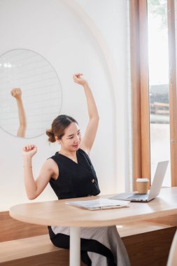 Genç bir kadın, modern bir ev ofisinde dizüstü bilgisayarla çalışırken doğal ışık ve bir fincan kahveyle esneme hareketleri yapıyor..