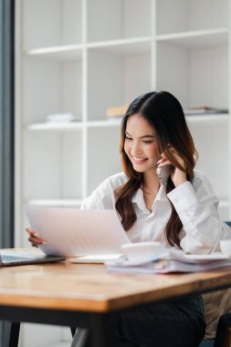 Bir kadın masada oturuyor ve cep telefonuyla konuşuyor. Gülümsüyor ve sohbetinden zevk alıyor. Masada bir sürü kağıt ve dizüstü bilgisayar var.
