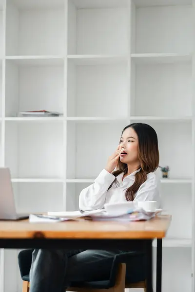 stock image A young woman yawning at her desk in a modern office setting, wearing a casual white shirt, working on a laptop, with white shelves in the background.