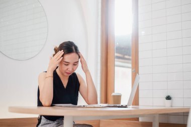Asyalı kadın evde çalışırken stres yaşıyor, minimalist bir masada dizüstü bilgisayar ve kahveyle oturuyor..