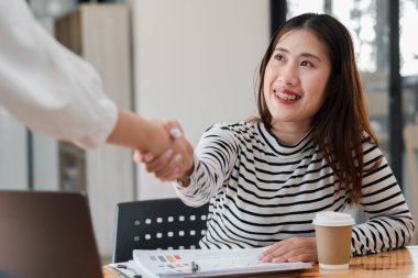 İş ortamında bir kadın başka bir kadınla tokalaşıyor. Soldaki kadın beyaz gömlek giyiyor ve sağdaki kadın da çizgili gömlek giyiyor.