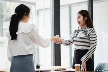 İş ortamında tokalaşan iki kadın. Bir kadın beyaz gömlek giyiyor, diğeri de çizgili gömlek.