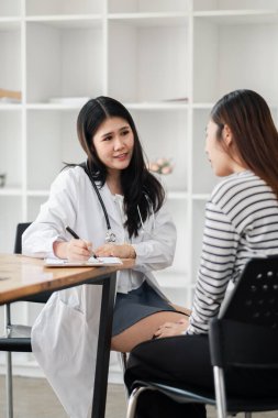 Bir kadın doktor bir kadın hastayla konuşuyor. Hasta çizgili bir gömlek giyiyor. Doktor bir kağıt parçasına bir şeyler yazıyor.