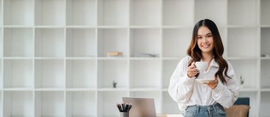 Gülümseyen genç bir kadın, arka planda minimalist beyaz bir rafla modern ofis ortamında elinde bir fincan kahve tutuyor..