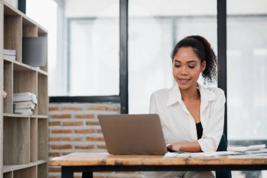 Modern bir ofiste dizüstü bilgisayarda çalışan genç bir kadın, tuğla duvar ve geniş pencereli, verimli bir çalışma ortamı sergiliyor..