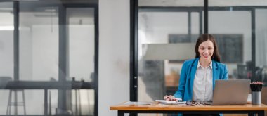 Mavi ceketli gülümseyen iş kadını, cam duvarlı ve minimalist dekorlu çağdaş bir ofiste dizüstü bilgisayarla çalışıyor..