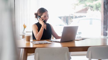 Bir kadın elinde dizüstü bilgisayarla bir masada otururken cep telefonuyla konuşuyor. Gülümsüyor ve sohbetinden zevk alıyor. Sahne rahat ve rahat bir atmosferi gösteriyor.