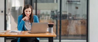 Bir kadın elinde dizüstü bilgisayar ve bir fincan kahveyle masada oturuyor. O, gözlük ve mavi bir ceket giyiyor. Verimlilik ve odaklanma kavramı, kadın büyük ihtimalle dizüstü bilgisayarı üzerinde çalışıyor.