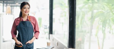 Tezgahın üzerinde kahve fincanı ve tabletle modern bir kafede cam kenarında duran önlüklü gülümseyen kadın barista..