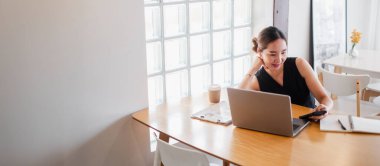 Modern bir ofis ortamında dizüstü bilgisayarıyla çalışan genç bir kadın. Masasında kahve fincanı ve not defteri var..