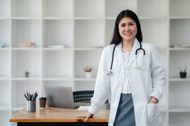 Beyaz laboratuvar önlüklü bir kadın elinde laptop ve bardakla bir masanın önünde duruyor. Gülümsüyor ve bir doktor.