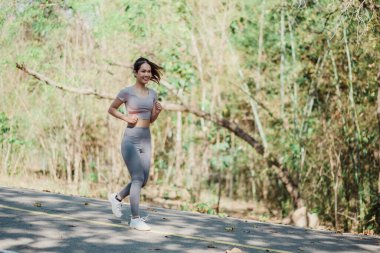 Orman parkında koşan genç bir kadın, atletik kıyafetler giyiyor ve gülümsüyor, güneşli bir günde açık havada egzersiz yapıyor..